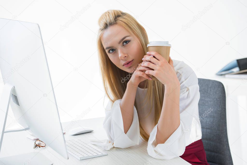 beautiful young businesswoman holding disposable coffee cup and looking at camera in office