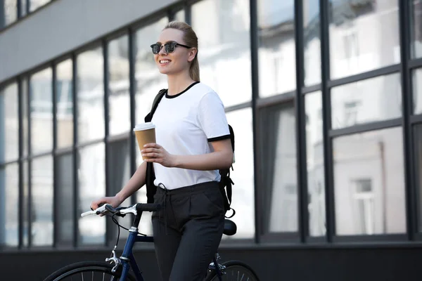 笑みを浮かべて美しいブロンドの女の子紙コップを押しながら路上自転車の側に立って — ストック写真