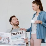 Marido leyendo el periódico de viaje mientras esposa con café de pie cerca
