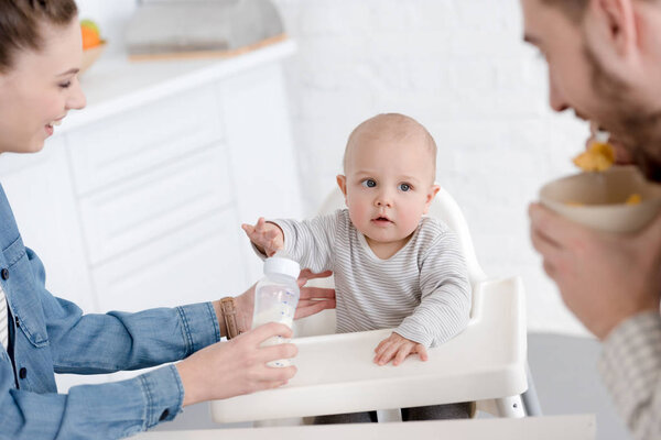 parents feeding baby boy with milk in baby bottle on kitchen