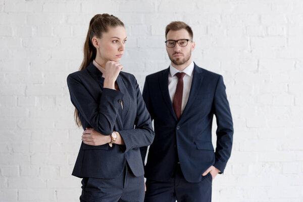 уверенный бизнесмен и задумчивая деловая женщина позируют возле белой стены
