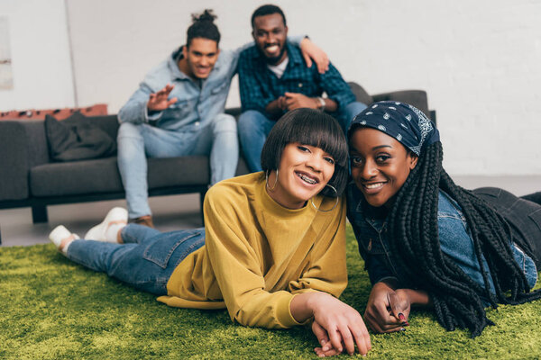 young smiling multiethnic women laying on rug and two male friends sitting behind