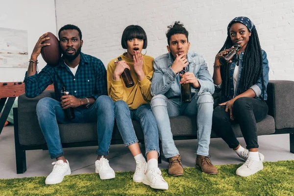 multicultural group of friends sitting on couch with beer bottles and watching american football match