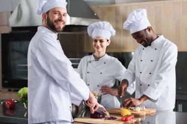 Multiracial team of cooks smiling while cutting ingredients by kitchen table clipart