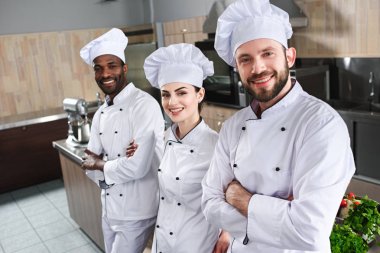Multiracial team of cooks looking at camera by cooking table clipart