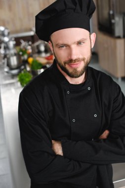 handsome chef standing with crossed arms and looking away at restaurant kitchen clipart