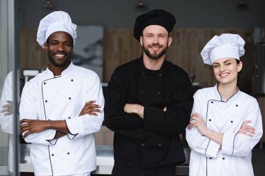 smiling multicultural chefs standing with crossed arms and looking at camera at restaurant kitchen
