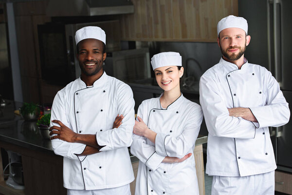smiling multicultural chefs standing with crossed arms at restaurant kitchen