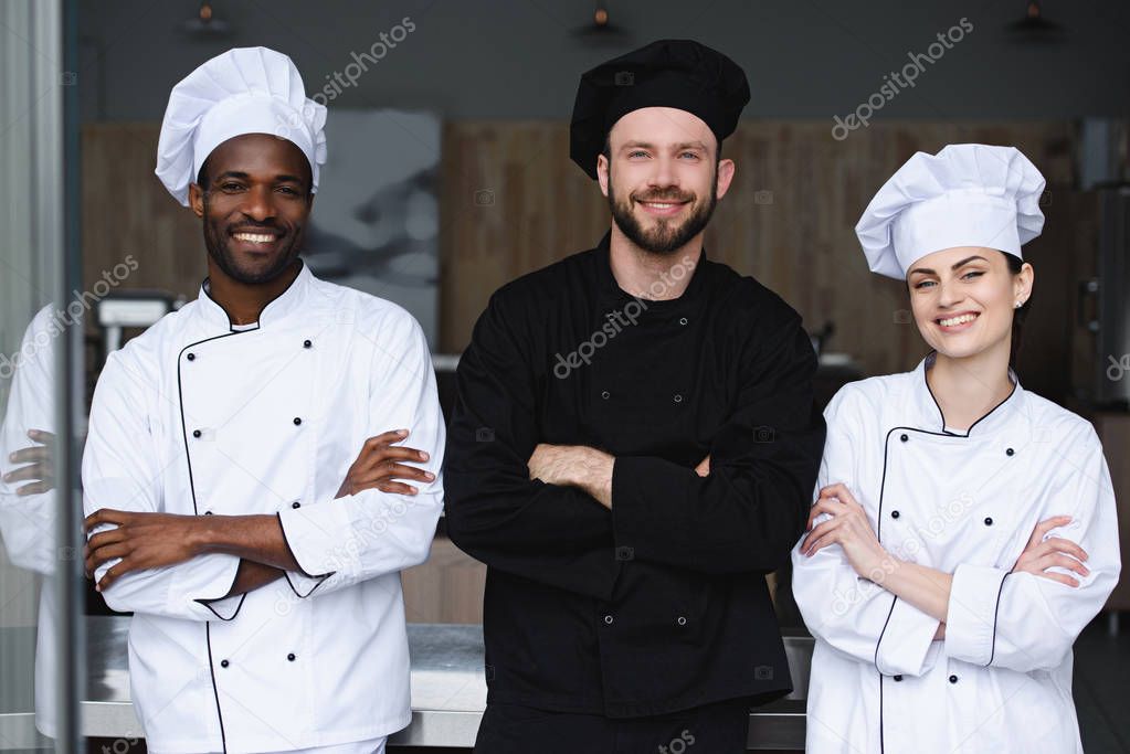 smiling multicultural chefs standing with crossed arms and looking at camera at restaurant kitchen