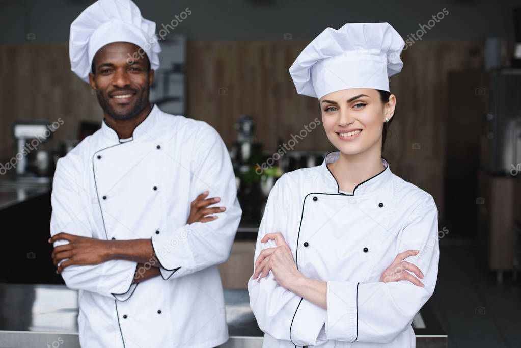 multicultural chefs standing with crossed arms and looking at camera at restaurant kitchen