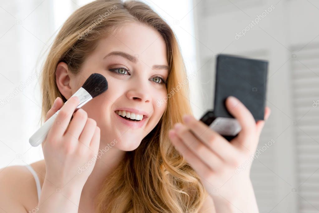 attractive smiling girl applying compact powder with brush and looking at mirror 