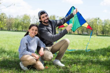 happy father and daughter sitting with kite on meadow in park clipart