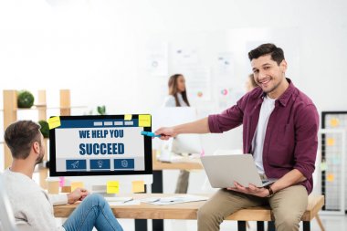 businessman pointing at we help you succeed inscription on computer screen while working at workplace with colleague in office clipart