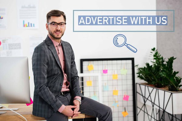 portrait of young businessman in suit and eyeglasses sitting on table in office, advertise with us inscription