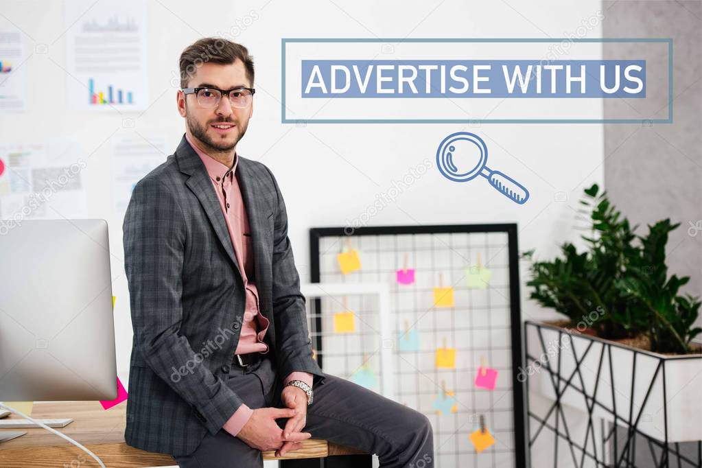 portrait of young businessman in suit and eyeglasses sitting on table in office, advertise with us inscription