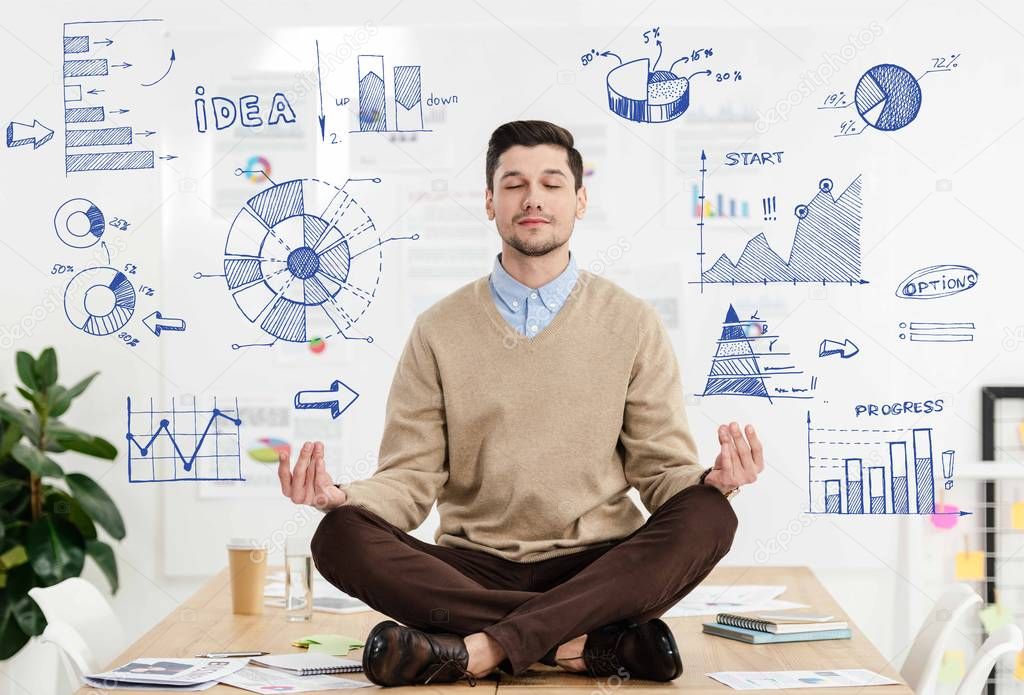 relaxed businessman with eyes closed sitting in lotus position on table in office with business graphs around