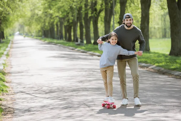 公園でスケート ボードに乗って彼女の娘の手を繋いでいる笑顔の父 — ストック写真