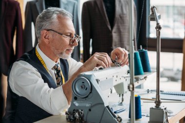 confident mature tailor working with modern sewing machine at workshop clipart