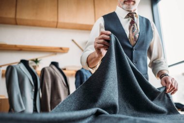 cropped shot of stylish mature tailor with grey cloth at sewing workshop clipart