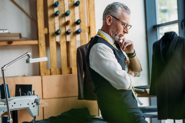 side view of handsome pensive tailor standing at sewing workshop