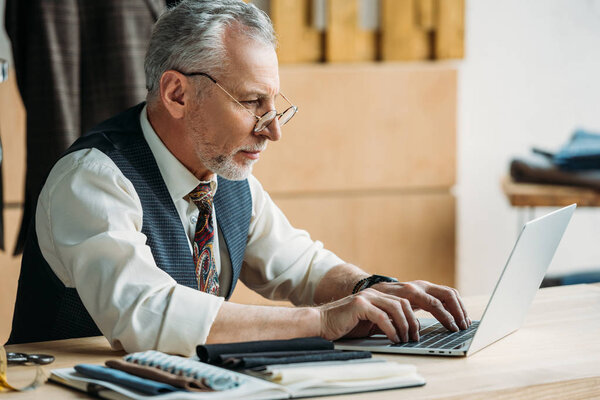 stylish mature tailor working with laptop at sewing workshop