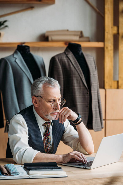 concentrated mature tailor working with laptop at sewing workshop