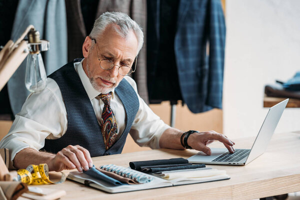 handsome mature tailor working with laptop and cloth samples at sewing workshop