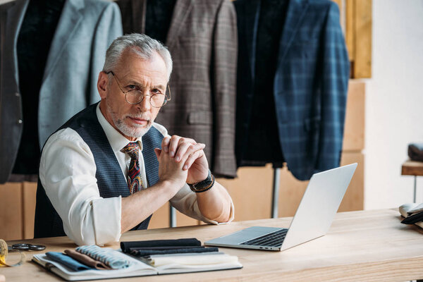 serious mature tailor with laptop on work desk at sewing workshop