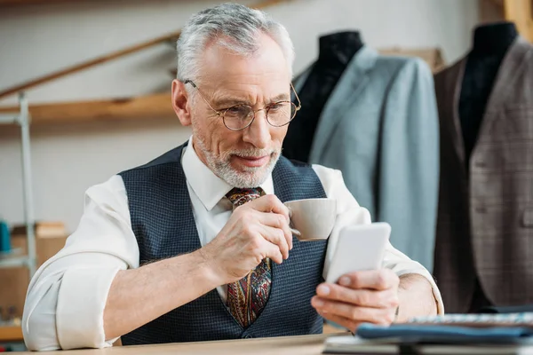 ハンサムな成熟した仕立てと縫製ワーク ショップでスマート フォンを使用してコーヒーを飲んで — ストック写真