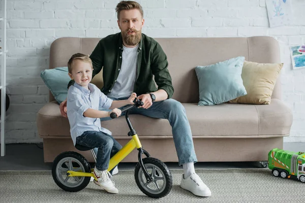 Niño Pequeño Bicicleta Equilibrio Con Padre Barbudo Sentado Sofá Casa — Foto de Stock