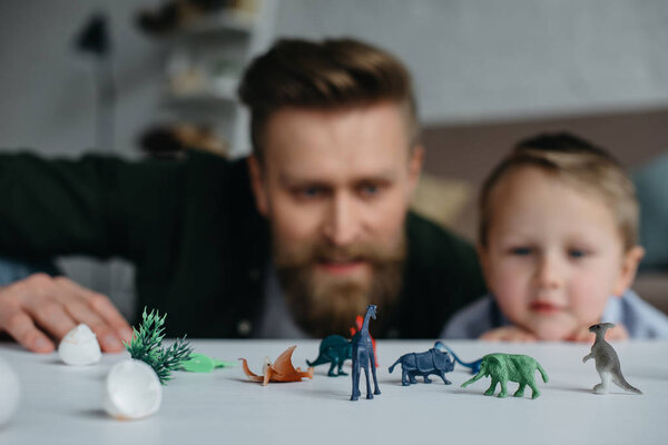 selective focus of father and cute little son looking at arranged toy dinosaurs on tabletop together at home