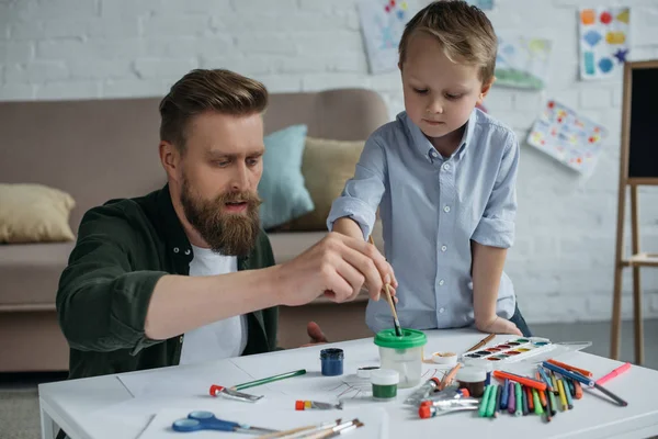 portrait of father and cute little son with paints and brushes drawing pictures together at home