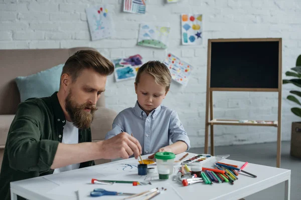 focused father and cute little son with paints and brushes drawing pictures together at home