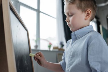 side view of little boy with piece of chalk drawing picture on blackboard at home clipart