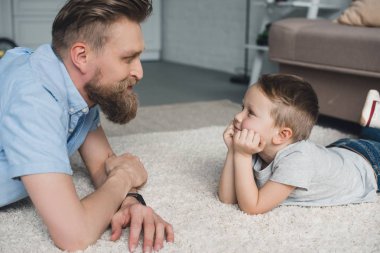 side view of bearded father and smiling son looking at each other at home clipart