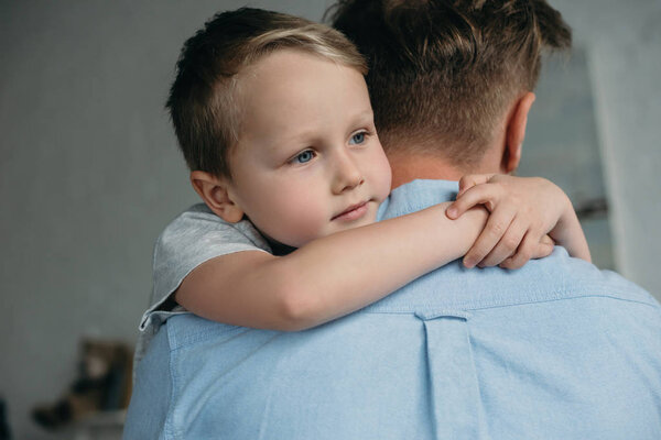 pensive little boy hugging father and looking away at home
