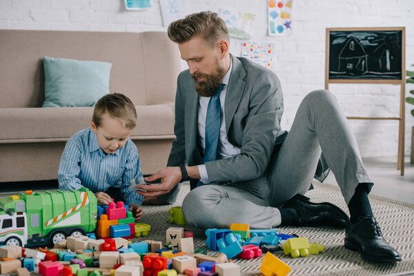 businessman in suit and little son playing with colorful blocks together on floor at home, work and life balance concept