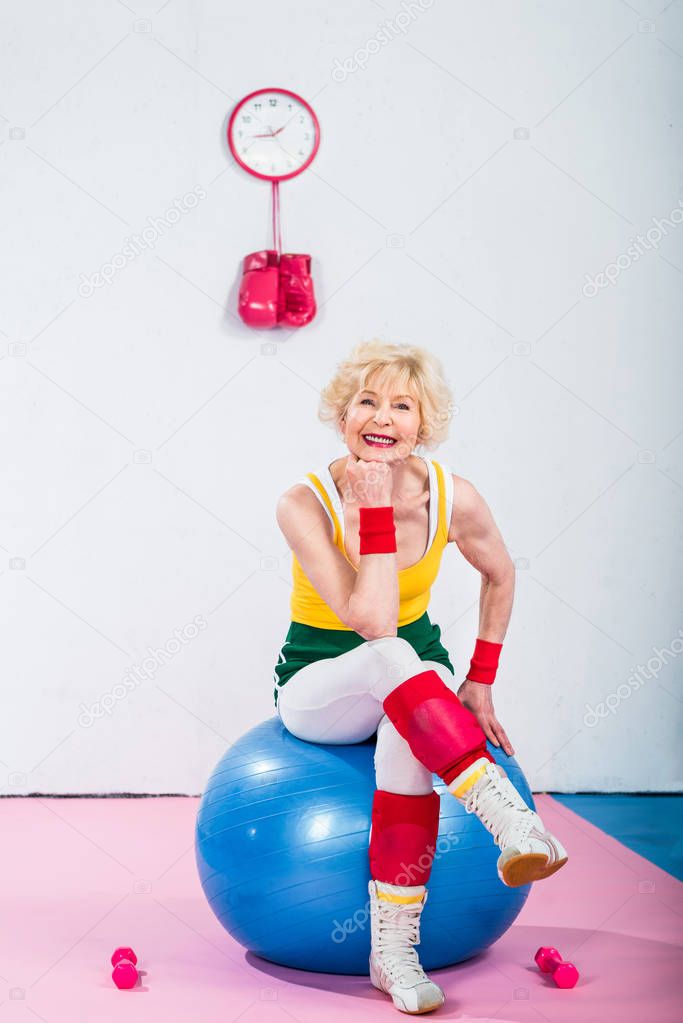happy sporty senior woman sitting on fitness ball and smiling at camera