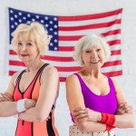 Belle donne anziane sportive in piedi con le braccia incrociate e sorridenti alla fotocamera contro la bandiera americana