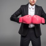 Vista recortada de una fuerte empresaria posando en guantes de boxeo, aislada en gris