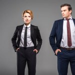 Serious businesswoman and businessman posing with hands in pockets, isolated on grey