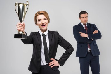 excited businesswoman with award and upset businessman behind, isolated on grey clipart