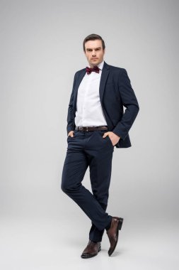 handsome man posing in tuxedo with hands in pockets, isolated on grey clipart