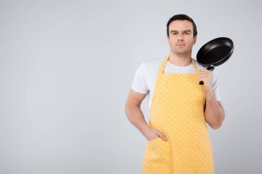 male householder in yellow apron holding frying pan, isolated on grey clipart
