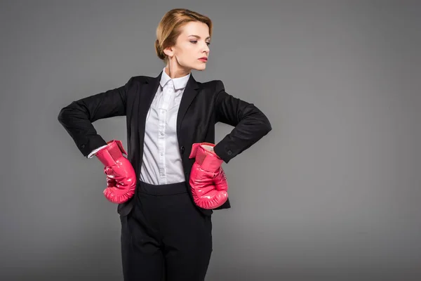 strong businesswoman posing in suit and pink boxing gloves, isolated on grey
