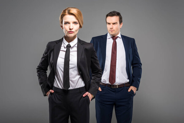 confident businesswoman and businessman in suits standing with hands in pockets, isolated on grey