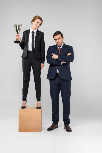 счастливая деловая женщина с кубком трофеев, стоящая на пьедестале почета, расстроенный бизнесмен, стоящий рядом, изолированный на сером
