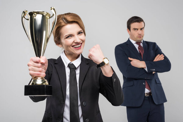 excited businesswoman with trophy cup and upset businessman behind, isolated on grey
