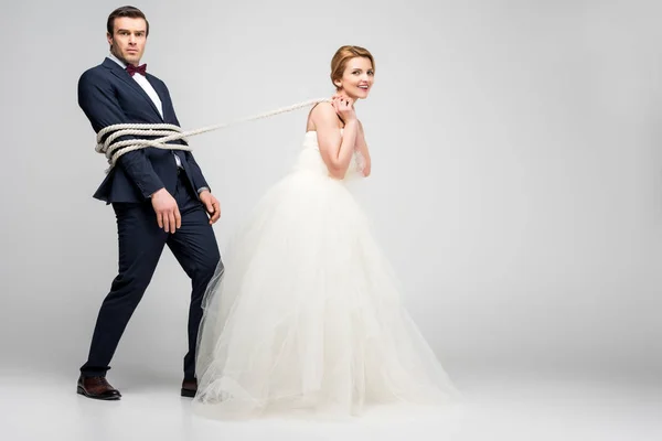 Schöne Braut Zieht Bräutigam Mit Seil Gebunden Isoliert Auf Grau — Stockfoto