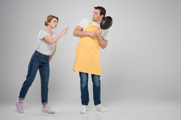 angry wife and frightened husband in apron with frying pan, feminism concept, isolated on grey
